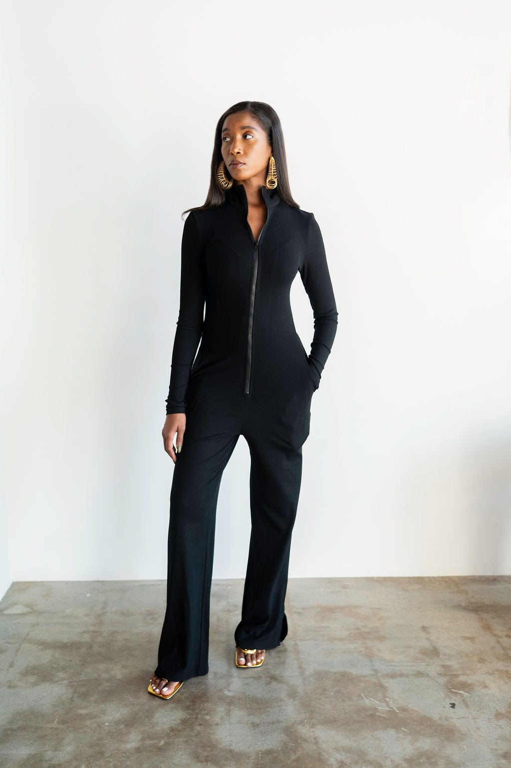 Woman wearing a black jumpsuit standing against a white wall.
