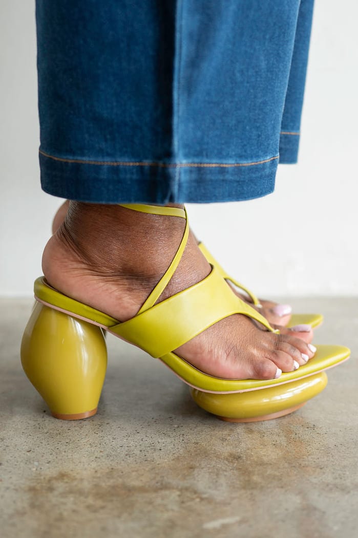 A close-up of a person's foot wearing a bright yellow strappy sandal with a chunky block heel, paired with blue jeans, shown against a neutral indoor background.
