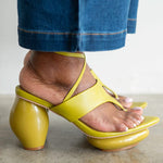 A close-up of a person's foot wearing a bright yellow strappy sandal with a chunky block heel, paired with blue jeans, shown against a neutral indoor background.
