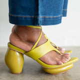 A close-up of a person's foot wearing a bright yellow strappy sandal with a chunky block heel, paired with blue jeans, shown against a neutral indoor background.