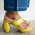 A close-up of a person's foot wearing a bright yellow strappy sandal with a chunky block heel, paired with blue jeans, shown against a neutral indoor background.