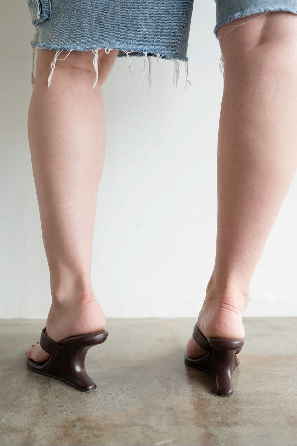 A person shown from the back wearing light blue frayed-hem denim shorts and dark brown heeled slide sandals with a low block heel, standing on a concrete floor against a plain light wall.