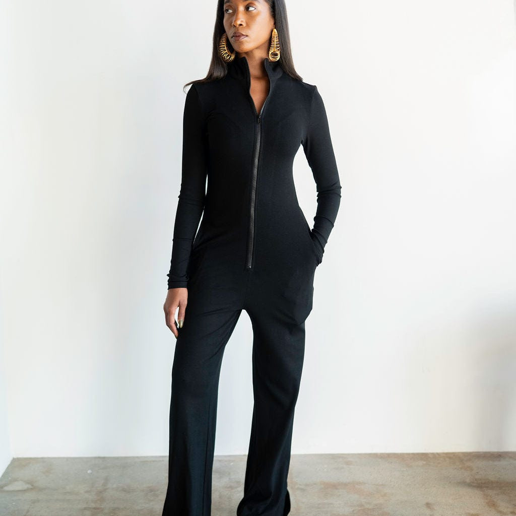 Woman wearing a black jumpsuit standing against a white wall.