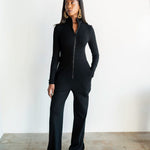 Woman wearing a black jumpsuit standing against a white wall.
