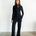 Woman wearing a black jumpsuit standing against a white wall.