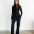 Woman wearing a black jumpsuit standing against a white wall.